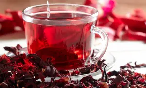 Ein Glas leuchtend roter Hibiskustee, umgeben von getrockneten Hibiskusblüten. Ist Hibiskustee gesund? Ja, er ist voller Antioxidantien.