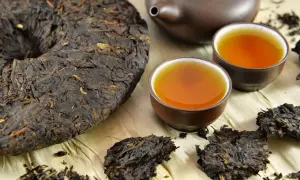 Ein runder Kuchen und lose Blätter von dunklem Pu-Erh-Tee neben zwei Tassen des bernsteinfarbenen Getränks und einer Teekanne.