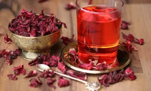 Getränk aus Malve: getrocknete Blüten in Schale, ein Glas roter Tee und eine Löffel auf Holztisch.