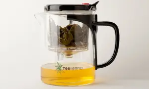 Glas-Teekanne mit schwarzem Deckel und Griff, gefüllt mit goldenem Tee und grünen Teeblättern. Perfekt, um Tee zu servieren.