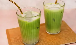 Zwei Gläser mit erfrischendem Matcha Hafermilch-Eisgetränk, serviert auf einem Holztablett. Goldene Strohhalme ragen aus den Getränken.