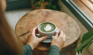Hände halten eine Tasse cremigen Matcha Latte mit Latte Art auf einem rustikalen Holztisch. Eine gesunde Alternative zu Kaffee.