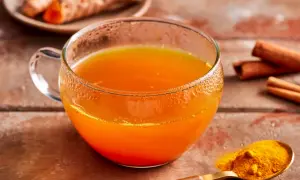 Ein Glas goldener Kurkuma Tee, daneben frische Kurkumawurzeln, Zimtstangen und ein Löffel Kurkumapulver. So einfach lässt sich Kurkuma Tee zubereiten.