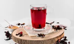 Ein Glas tiefroter Hibiskus-Tee, bereit zur Zubereitung. Getrocknete Hibiskusblüten und Honig sind daneben arrangiert.