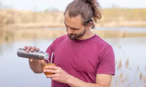 Mann mit Dutt und Bart gießt heißes Wasser aus Thermoskanne in Kalebasse für Mate oder Matcha.