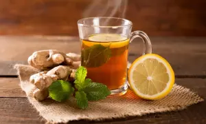 Heißer Tee mit Ingwer, Zitrone und Minze dampft in einer Tasse. Frische Ingwerwurzeln und eine Zitronenhälfte liegen daneben.