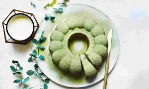 Ein köstlicher veganer Matcha-Kuchen, bestäubt mit Puderzucker, liegt auf einem Teller. Daneben ein Glas mit weißer Flüssigkeit und grünes Blattwerk.