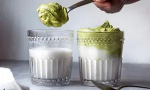 Ein Löffel mit cremigem Dalgona Matcha wird über ein Glas Milch gehalten. Daneben steht ein fertiges Glas Dalgona Matcha.