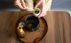 Vorbereitung von Matcha für die Schwangerschaft: Hände schöpfen grünes Pulver in eine Schale mit heißem Wasser. Ein Chasen (Bambusbesen) liegt daneben.