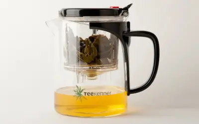 Glas-Teekanne mit schwarzem Deckel und Griff, gefüllt mit goldenem Tee und grünen Teeblättern. Perfekt, um Tee zu servieren.