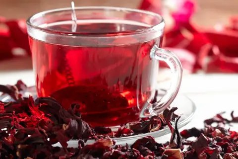 Ein Glas leuchtend roter Hibiskustee, umgeben von getrockneten Hibiskusblüten. Ist Hibiskustee gesund? Ja, er ist voller Antioxidantien.