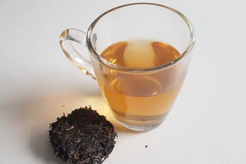 Ein Glas goldbrauner Pu-erh-Tee neben trockenen Teeblättern. Der Tee liefert sanftes Koffein für einen belebenden Start.