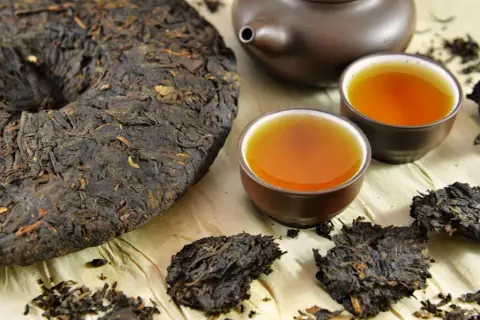 Ein runder Kuchen und lose Blätter von dunklem Pu-Erh-Tee neben zwei Tassen des bernsteinfarbenen Getränks und einer Teekanne.