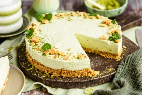 Ein cremiger Matcha Cheesecake mit Keksboden, garniert mit Minze und zerbröselten Keksen. Ein Stück wurde bereits angeschnitten.