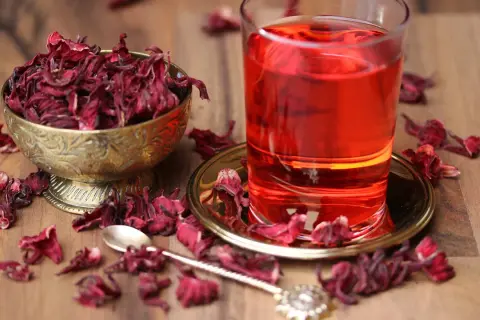 Getränk aus Malve: getrocknete Blüten in Schale, ein Glas roter Tee und eine Löffel auf Holztisch.