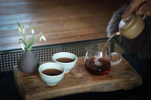 Ein sanfter Strahl dunklen Tees fließt aus einer kleinen Kanne in ein Glasgefäß. Die Pu-erh-Tee-Wirkung entfaltet sich in der ruhigen Atmosphäre, begleitet von Schneeglöckchen.