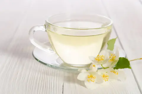 Eine Tasse klarer, heller Flüssigkeit, die an weißen Tee erinnert, mit duftenden Jasminblüten daneben.