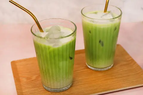 Zwei Gläser mit erfrischendem Matcha Hafermilch-Eisgetränk, serviert auf einem Holztablett. Goldene Strohhalme ragen aus den Getränken.