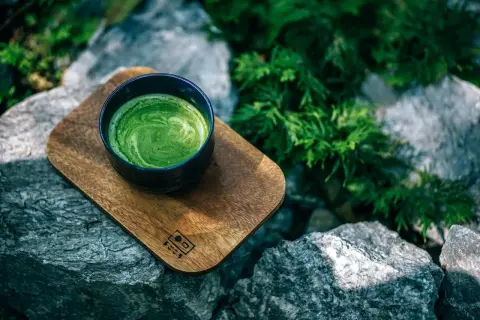 Eine Schale cremiger Matcha-Tee auf einem Holzbrett, umgeben von Steinen und Grün. Perfekt für eine ruhige Matcha-Stillzeit.