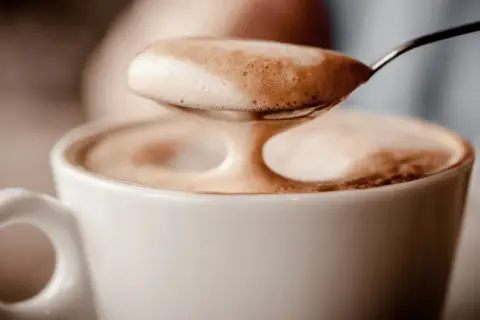 Ein Löffel voller Milchschaum über einer Tasse Kaffee. Der Koffein-Kick für den Magen.
