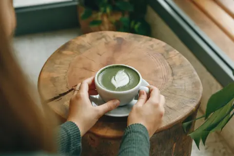 Hände halten eine Tasse cremigen Matcha Latte mit Latte Art auf einem rustikalen Holztisch. Eine gesunde Alternative zu Kaffee.