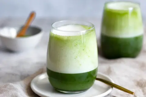 Zwei Gläser mit geschichtetem Matcha-Latte, die die perfekte Matcha-Zubereitung zeigen. Ein Löffel liegt daneben.