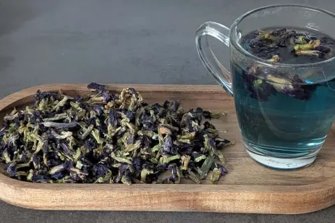 Getränk aus Schmetterlingserbse Tee, dessen Wirkung durch die tiefblaue Farbe und die getrockneten Blüten auf dem Holztablett angedeutet wird.