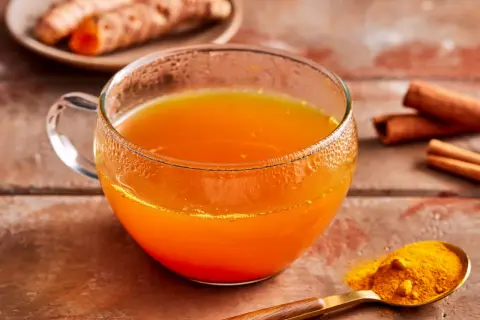 Ein Glas goldener Kurkuma Tee, daneben frische Kurkumawurzeln, Zimtstangen und ein Löffel Kurkumapulver. So einfach lässt sich Kurkuma Tee zubereiten.