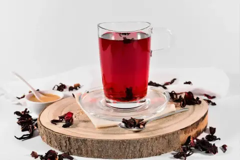 Ein Glas tiefroter Hibiskus-Tee, bereit zur Zubereitung. Getrocknete Hibiskusblüten und Honig sind daneben arrangiert.