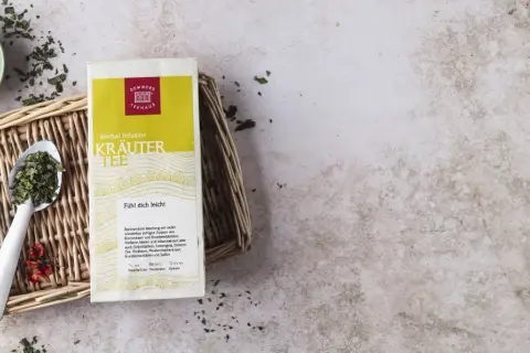 Kräutertee-Packung mit Löffel und Schale voller Kräuter. Die tee temperatur tabelle gibt Hinweise zur Zubereitung.