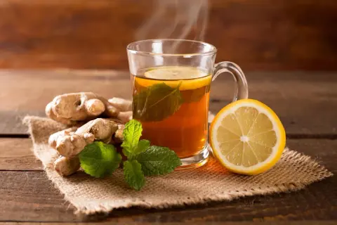 Heißer Tee mit Ingwer, Zitrone und Minze dampft in einer Tasse. Frische Ingwerwurzeln und eine Zitronenhälfte liegen daneben.