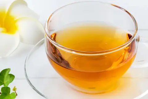 Klarer schwarzer Tee in einer Glas Tasse, beruhigend für den Magen, neben einer weißen Blüte und grünen Blättern.