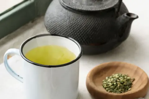 Dampfender Genmaicha in einer Tasse, daneben eine Teekanne und eine Schale mit geröstetem Reis. Die Genmaicha Zubereitung ist im Gange.