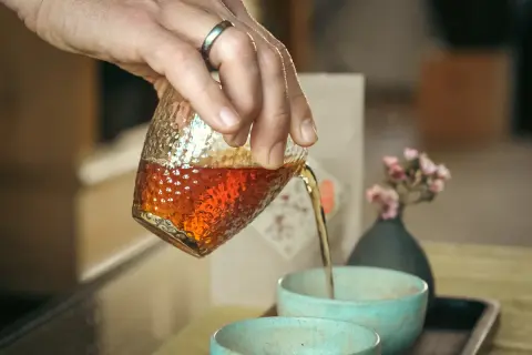 Hand gießt schwarzen Japaner Tee in eine Schale. Ein kleiner Strauß rosa Blüten steht daneben.