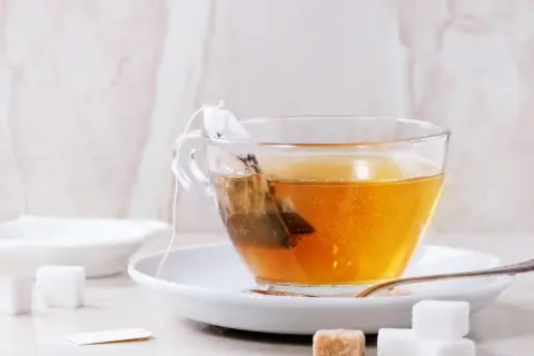 Eine Tasse Tee mit einem Teebeutel, der aussieht, als wäre der abgelaufene Tee schon lange im Wasser. Zuckerwürfel liegen daneben.