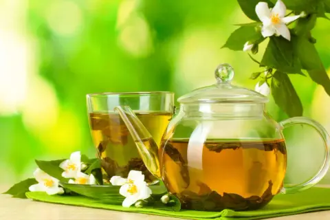 Glas-Teekanne und Tasse mit goldenem Jasmin Tee, umgeben von frischen Jasminblüten. Perfekt für die Jasmin Tee Zubereitung.