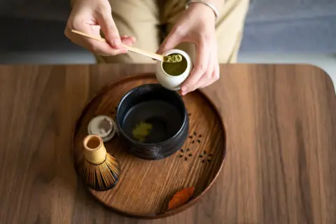Vorbereitung von Matcha für die Schwangerschaft: Hände schöpfen grünes Pulver in eine Schale mit heißem Wasser. Ein Chasen (Bambusbesen) liegt daneben.