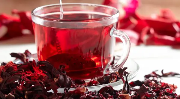 Ein Glas leuchtend roter Hibiskustee, umgeben von getrockneten Hibiskusblüten. Ist Hibiskustee gesund? Ja, er ist voller Antioxidantien.