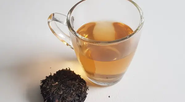 Ein Glas goldbrauner Pu-erh-Tee neben trockenen Teeblättern. Der Tee liefert sanftes Koffein für einen belebenden Start.