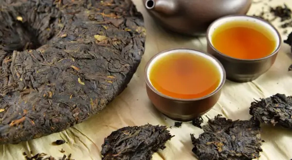 Ein runder Kuchen und lose Blätter von dunklem Pu-Erh-Tee neben zwei Tassen des bernsteinfarbenen Getränks und einer Teekanne.