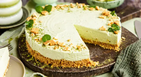 Ein cremiger Matcha Cheesecake mit Keksboden, garniert mit Minze und zerbröselten Keksen. Ein Stück wurde bereits angeschnitten.