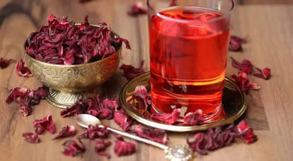 Getränk aus Malve: getrocknete Blüten in Schale, ein Glas roter Tee und eine Löffel auf Holztisch.