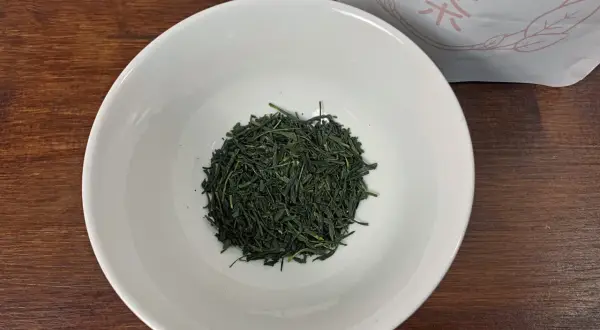 Trockene, dunkelgrüne Sencha-Teeblätter liegen in einer weißen Schale, bereit für die Zubereitung.