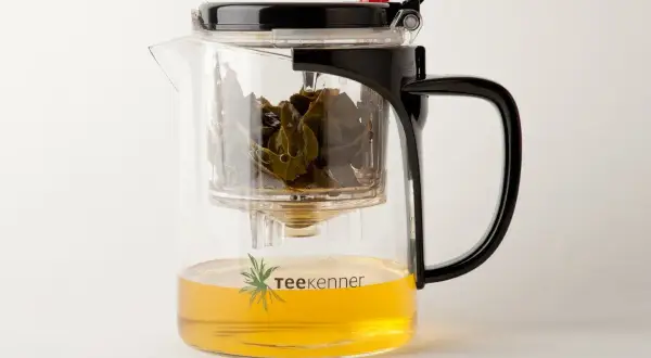 Glas-Teekanne mit schwarzem Deckel und Griff, gefüllt mit goldenem Tee und grünen Teeblättern. Perfekt, um Tee zu servieren.