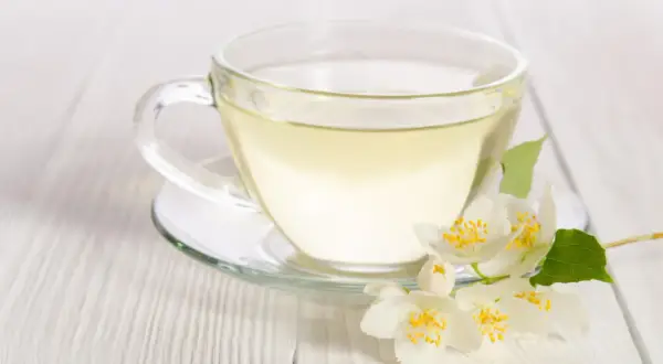 Eine Tasse klarer, heller Flüssigkeit, die an weißen Tee erinnert, mit duftenden Jasminblüten daneben.
