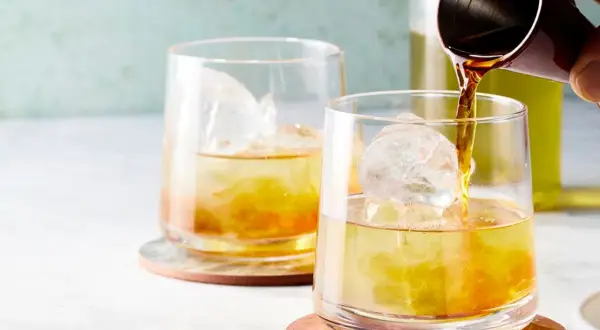 Zwei Gläser mit Eis und einem bernsteinfarbenen Getränk. Ein Jigger gießt Flüssigkeit in das vordere Glas. Ein erfrischender Gin mit Eistee.