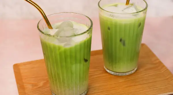 Zwei Gläser mit erfrischendem Matcha Hafermilch-Eisgetränk, serviert auf einem Holztablett. Goldene Strohhalme ragen aus den Getränken.