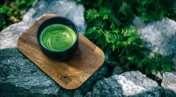 Eine Schale cremiger Matcha-Tee auf einem Holzbrett, umgeben von Steinen und Grün. Perfekt für eine ruhige Matcha-Stillzeit.