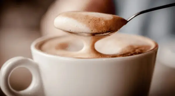Ein Löffel voller Milchschaum über einer Tasse Kaffee. Der Koffein-Kick für den Magen.
