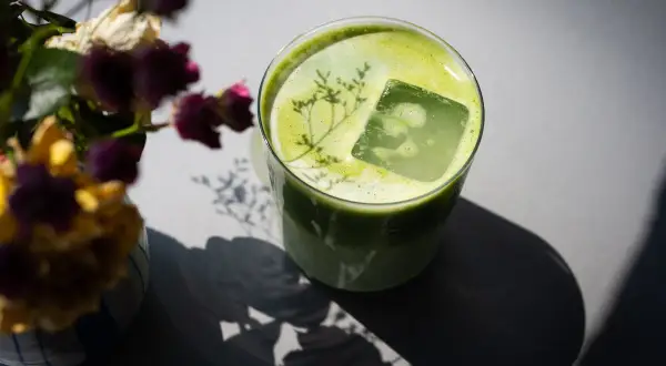 Ein Glas grüner Matcha mit Eiswürfel und einer Pflanze darauf. Neben dem Glas steht ein Strauß getrockneter Blumen. Mögliche matcha nebenwirkungen sind hier nicht ersichtlich.
